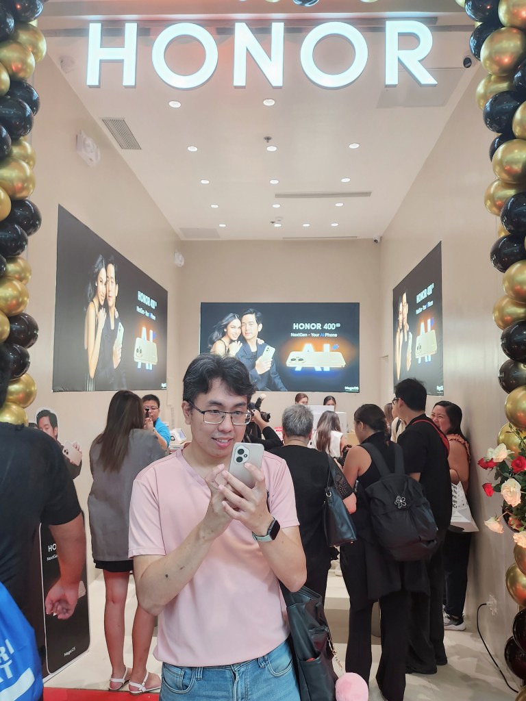 Author Bonne Zalzos holding a smartphone inside the HONOR store during the launch event of the HONOR 400 5G, surrounded by a crowd with displays featuring the smartphone.